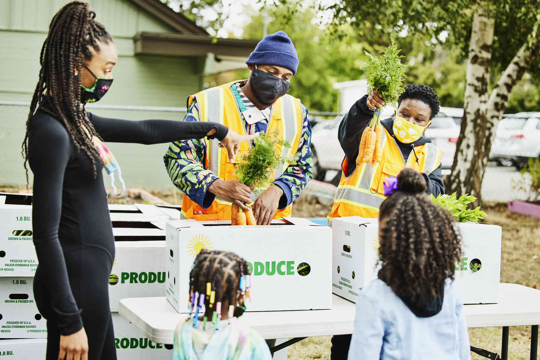 Two men wearing masks stand behind a table packing boxes of produce and handing them out to people waiting in a line next to the table.